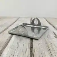 A square, hammered copper tray with a raised edge and two decorative leaf-shaped handles. The tray is sitting on a wooden surface.